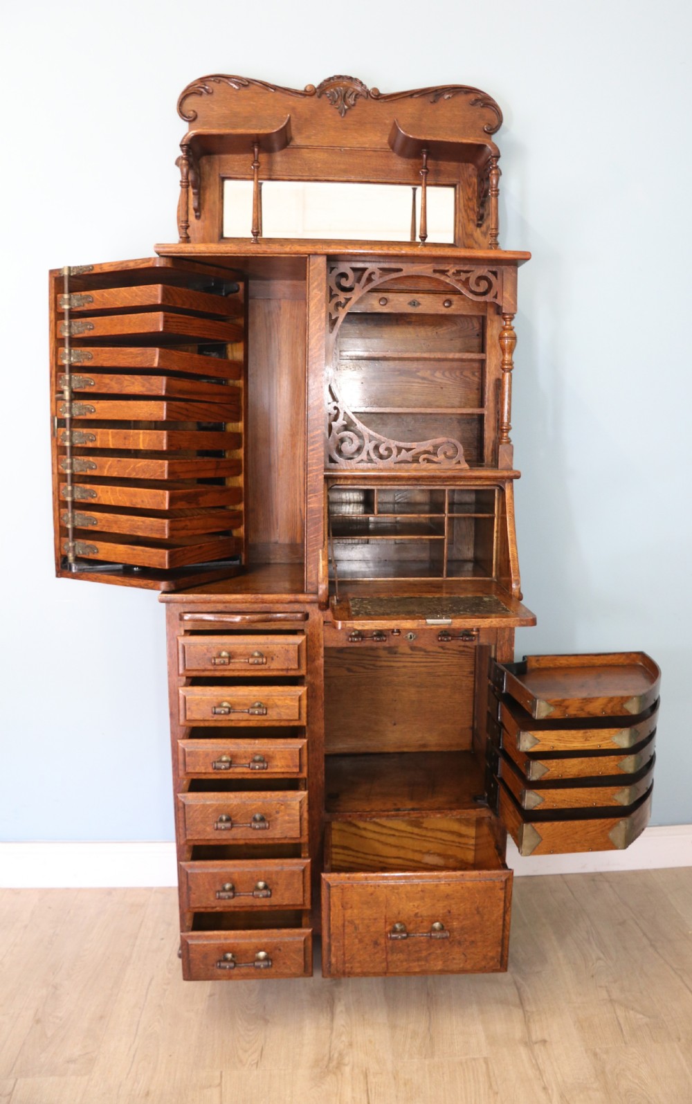 A late nineteenth-century oak combination dentist cabinet featuring a mirrored gallery top, carved foliate door, tambour section, and multiple small drawers. View V