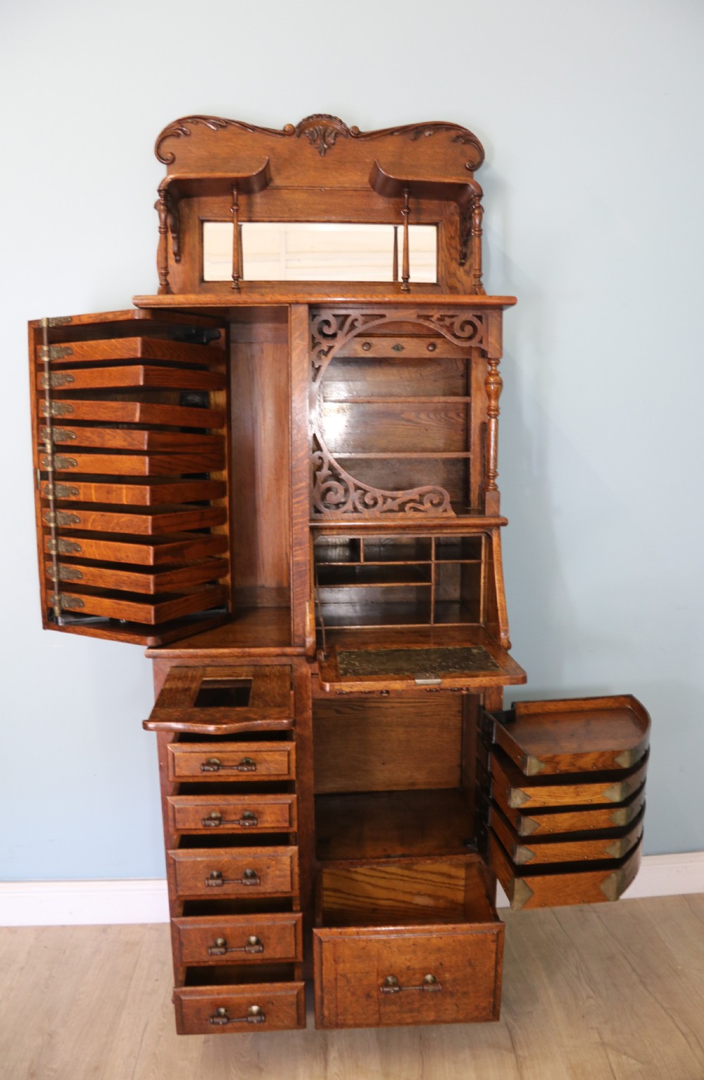 A late nineteenth-century oak combination dentist cabinet featuring a mirrored gallery top, carved foliate door, tambour section, and multiple small drawers. View VI