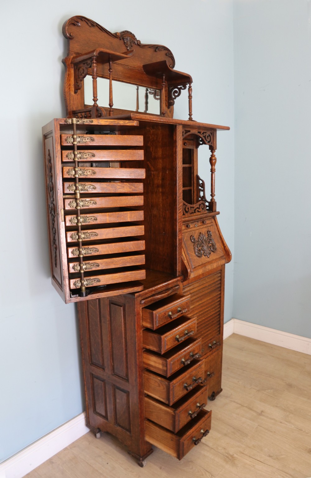 A late nineteenth-century oak combination dentist cabinet featuring a mirrored gallery top, carved foliate door, tambour section, and multiple small drawers. View VIII