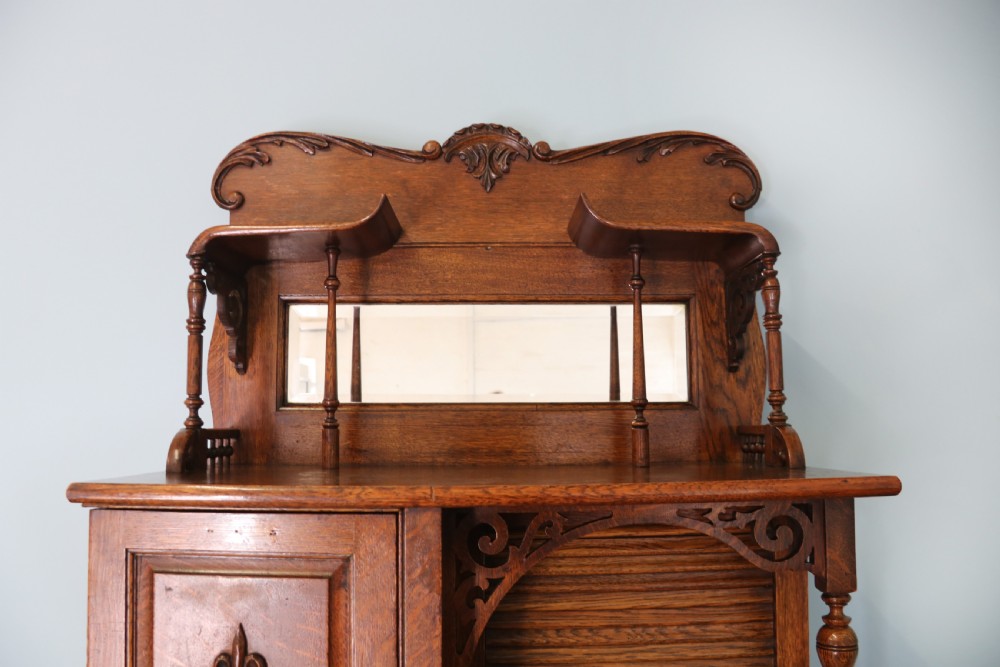 A late nineteenth-century oak combination dentist cabinet featuring a mirrored gallery top, carved foliate door, tambour section, and multiple small drawers. View X