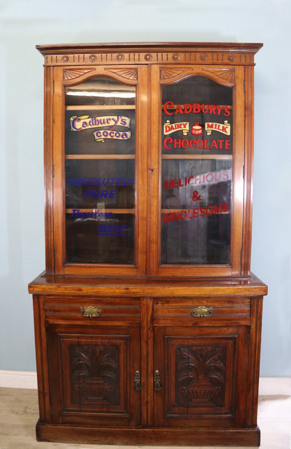 A two-tier dark walnut display cabinet featuring glazed upper doors decorated with vintage Cadburys advertising lettering above lower carved wooden cupboard doors.