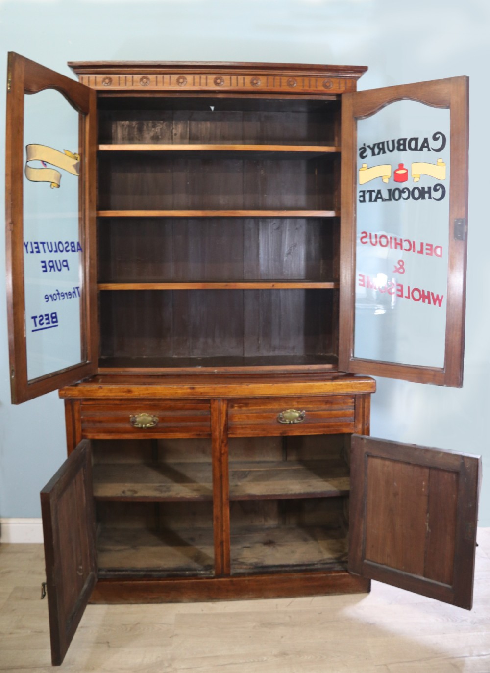 A two-tier dark walnut display cabinet featuring glazed upper doors decorated with vintage Cadburys advertising lettering above lower carved wooden cupboard doors. View II