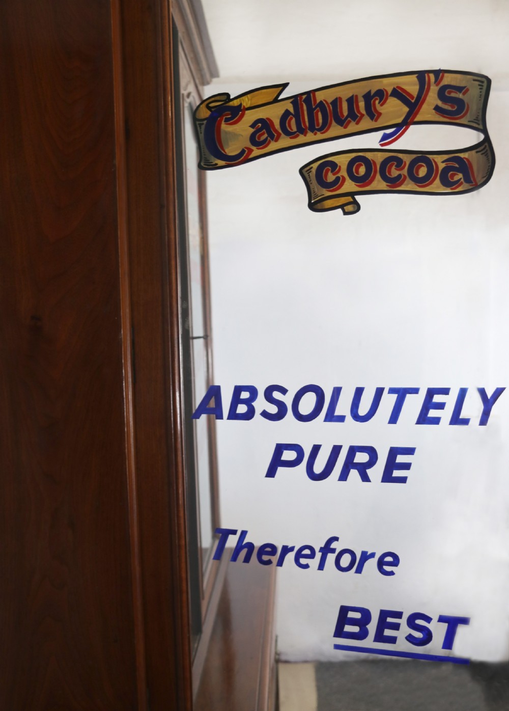 A two-tier dark walnut display cabinet featuring glazed upper doors decorated with vintage Cadburys advertising lettering above lower carved wooden cupboard doors. View IV