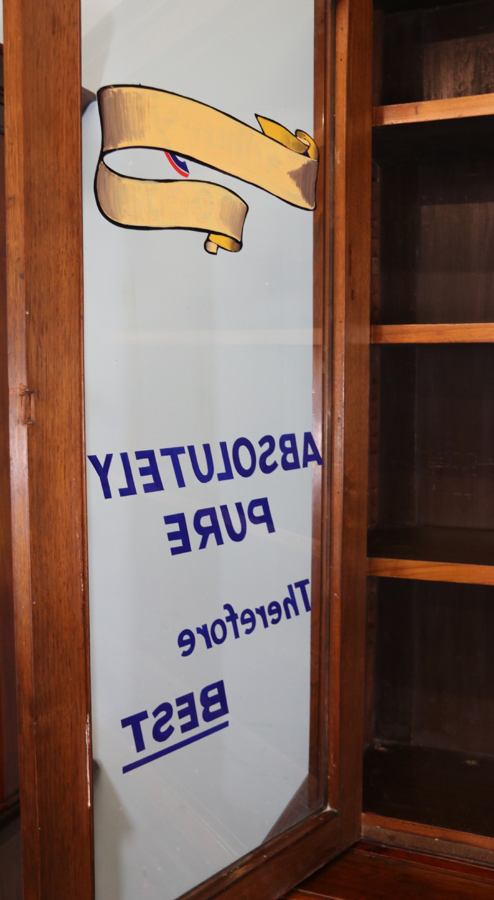 A two-tier dark walnut display cabinet featuring glazed upper doors decorated with vintage Cadburys advertising lettering above lower carved wooden cupboard doors. View V