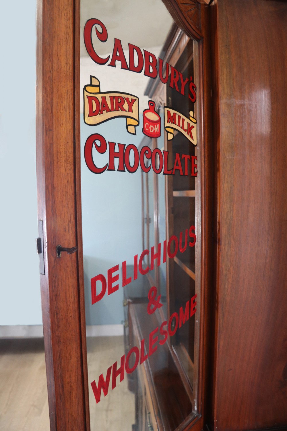 A two-tier dark walnut display cabinet featuring glazed upper doors decorated with vintage Cadburys advertising lettering above lower carved wooden cupboard doors. View VI
