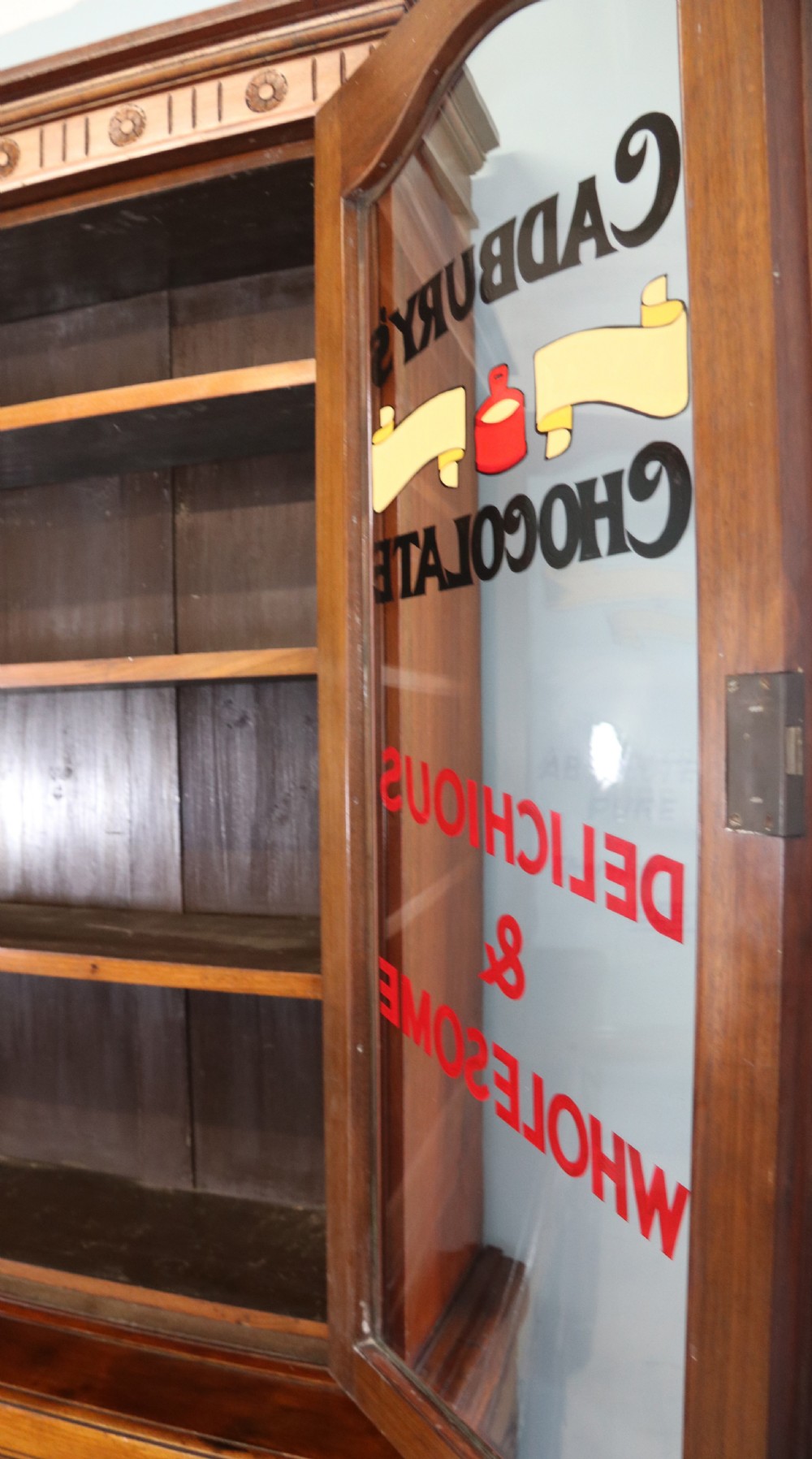 A two-tier dark walnut display cabinet featuring glazed upper doors decorated with vintage Cadburys advertising lettering above lower carved wooden cupboard doors. View VII