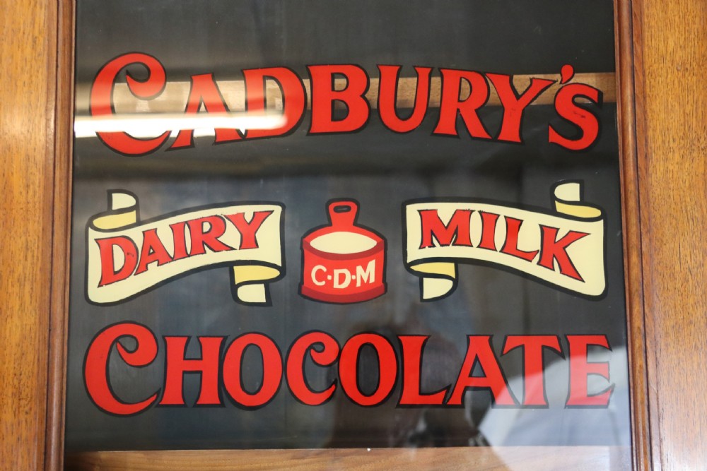 A two-tier dark walnut display cabinet featuring glazed upper doors decorated with vintage Cadburys advertising lettering above lower carved wooden cupboard doors. View XIII