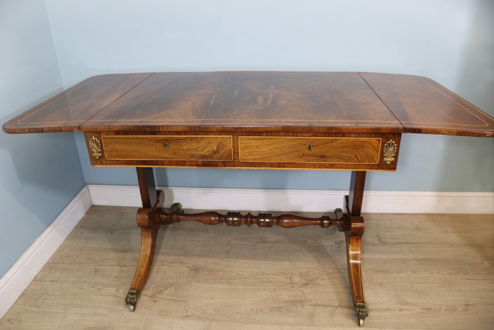 An early 19th-century Regency drop-leaf sofa table made of polished dark wood with two drawers, brass mounts, turned stretcher, and splayed legs with brass paw castors.