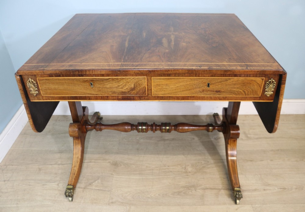 An early 19th-century Regency drop-leaf sofa table made of polished dark wood with two drawers, brass mounts, turned stretcher, and splayed legs with brass paw castors. View II