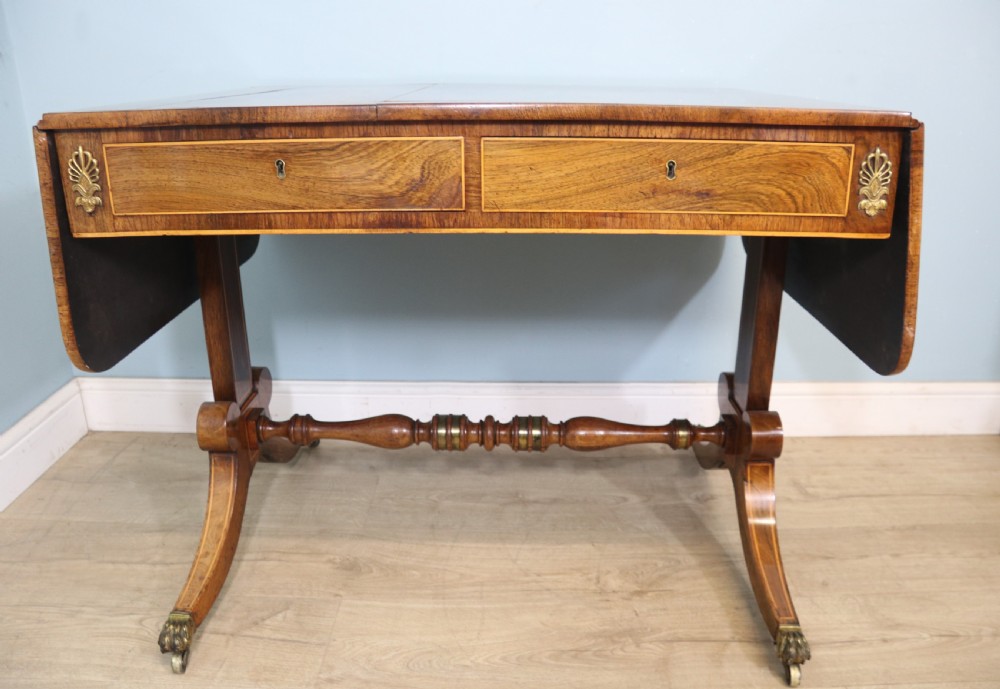 An early 19th-century Regency drop-leaf sofa table made of polished dark wood with two drawers, brass mounts, turned stretcher, and splayed legs with brass paw castors. View III
