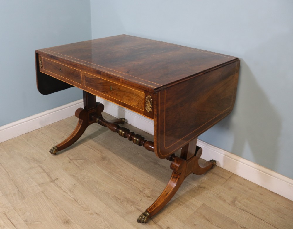 An early 19th-century Regency drop-leaf sofa table made of polished dark wood with two drawers, brass mounts, turned stretcher, and splayed legs with brass paw castors. View IV