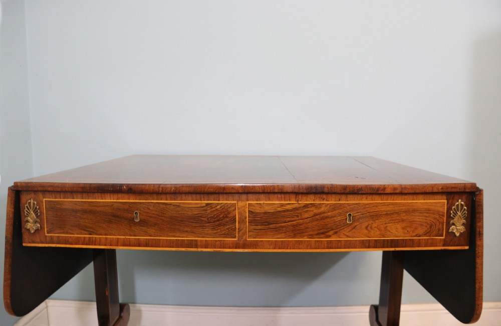 An early 19th-century Regency drop-leaf sofa table made of polished dark wood with two drawers, brass mounts, turned stretcher, and splayed legs with brass paw castors. View V