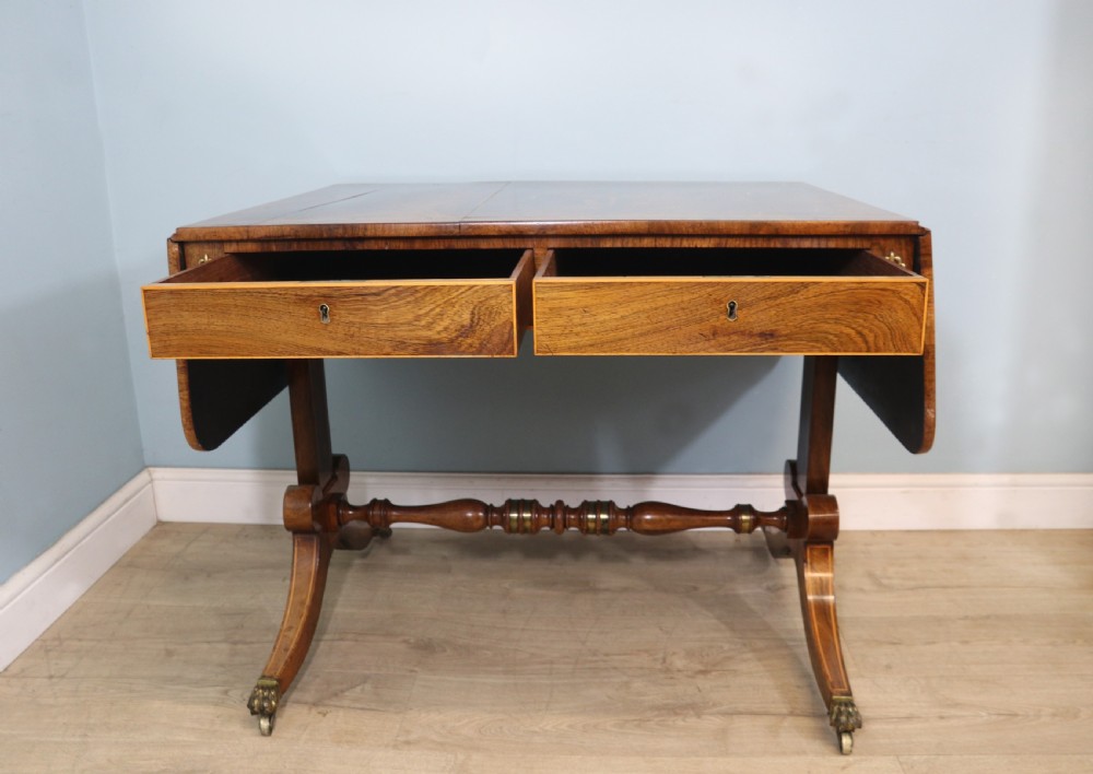 An early 19th-century Regency drop-leaf sofa table made of polished dark wood with two drawers, brass mounts, turned stretcher, and splayed legs with brass paw castors. View VI