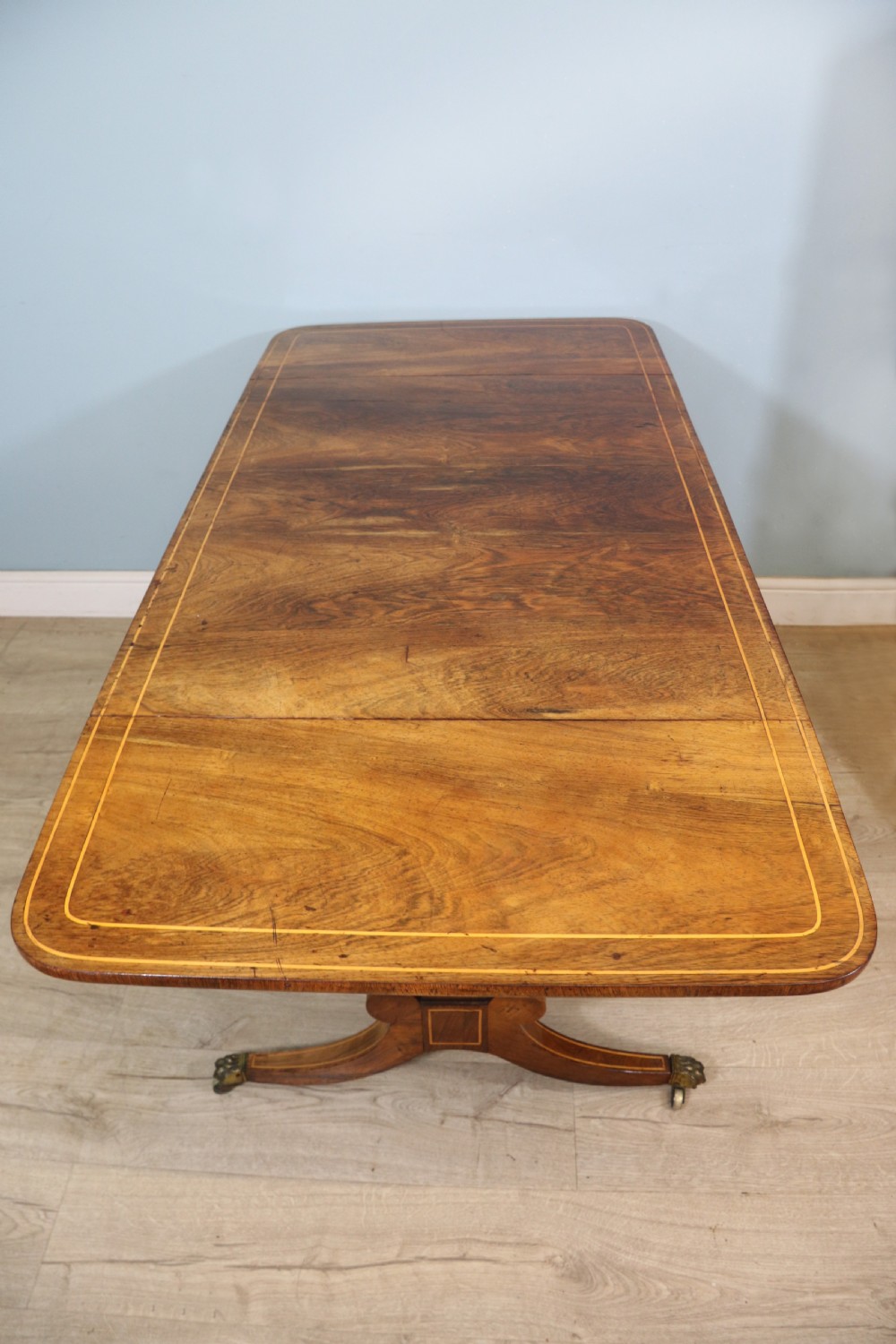 An early 19th-century Regency drop-leaf sofa table made of polished dark wood with two drawers, brass mounts, turned stretcher, and splayed legs with brass paw castors. View X