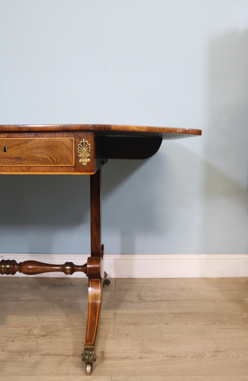 An early 19th-century Regency drop-leaf sofa table made of polished dark wood with two drawers, brass mounts, turned stretcher, and splayed legs with brass paw castors. View XI