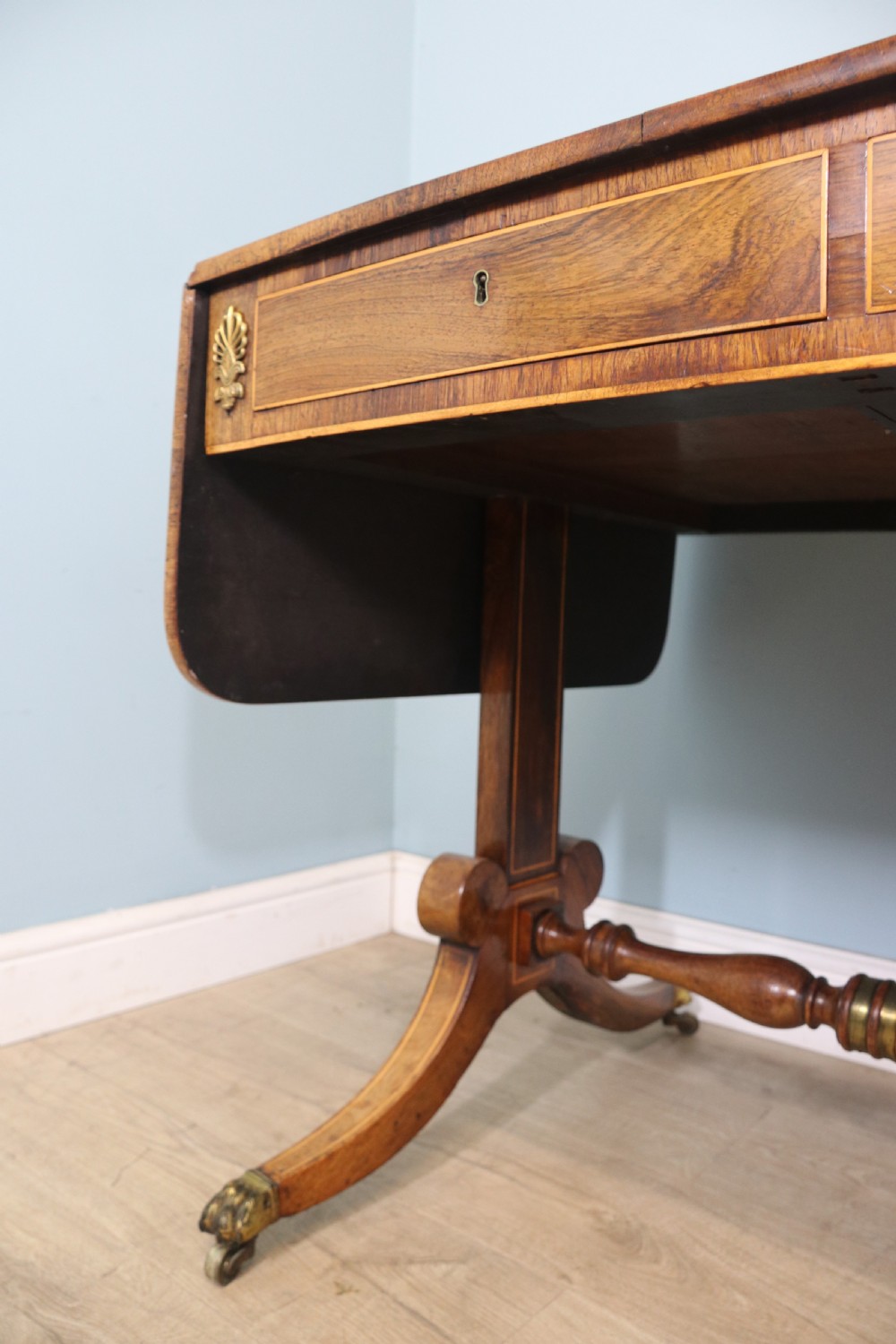 An early 19th-century Regency drop-leaf sofa table made of polished dark wood with two drawers, brass mounts, turned stretcher, and splayed legs with brass paw castors. View XIX