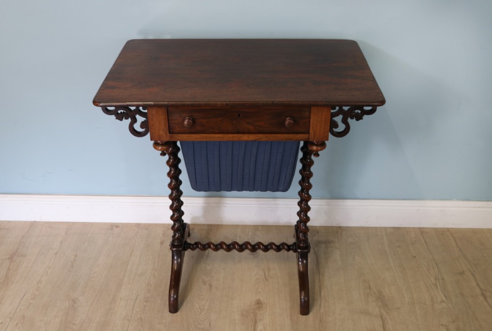 A 19th-century dark rosewood sewing table featuring barley twist legs, side fretwork brackets, a single drawer, and a blue fabric work basket below.