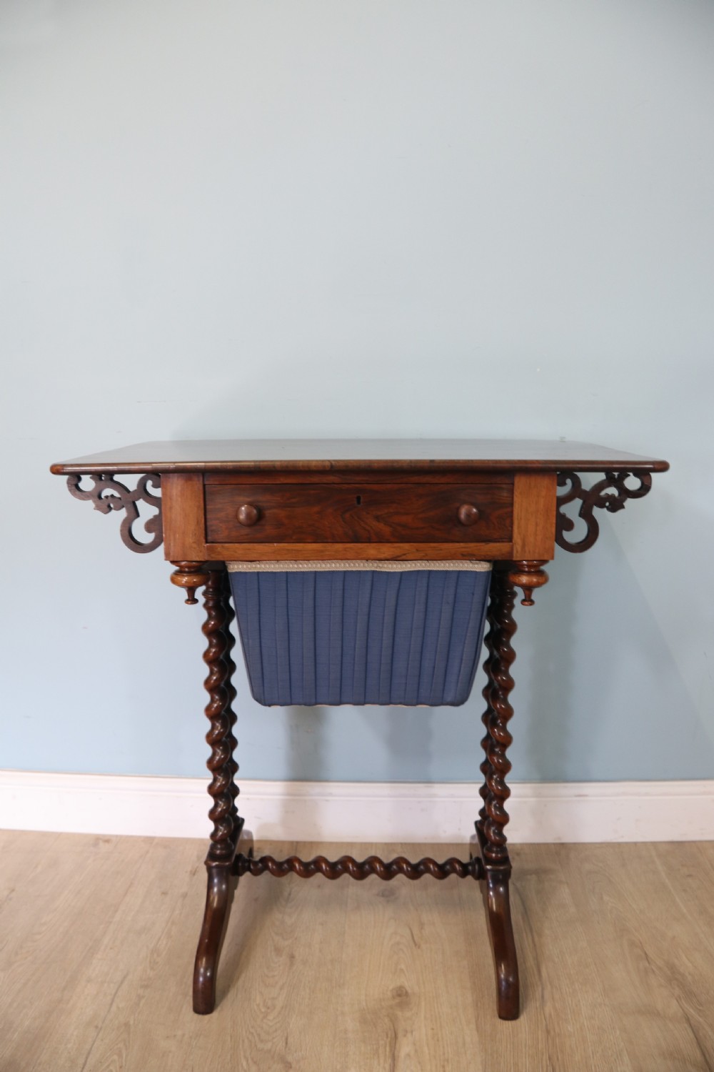 A 19th-century dark rosewood sewing table featuring barley twist legs, side fretwork brackets, a single drawer, and a blue fabric work basket below. View II