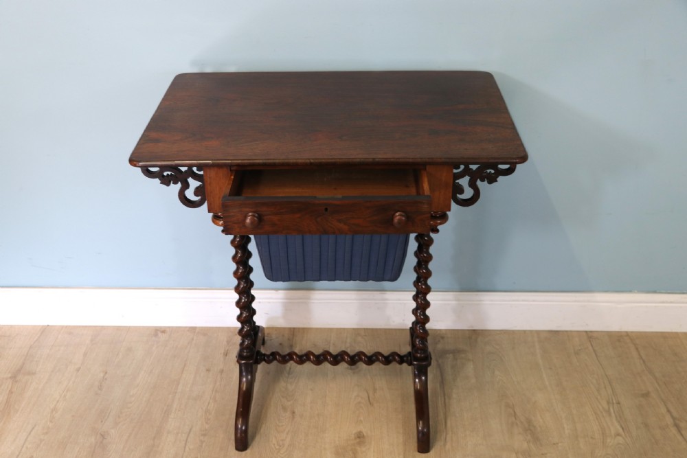 A 19th-century dark rosewood sewing table featuring barley twist legs, side fretwork brackets, a single drawer, and a blue fabric work basket below. View IV