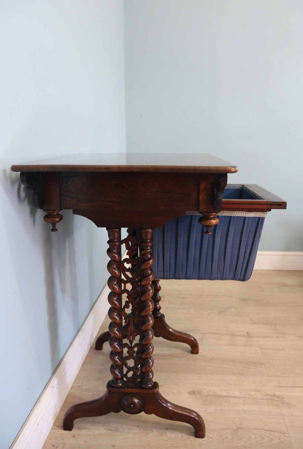 A 19th-century dark rosewood sewing table featuring barley twist legs, side fretwork brackets, a single drawer, and a blue fabric work basket below. View XVI