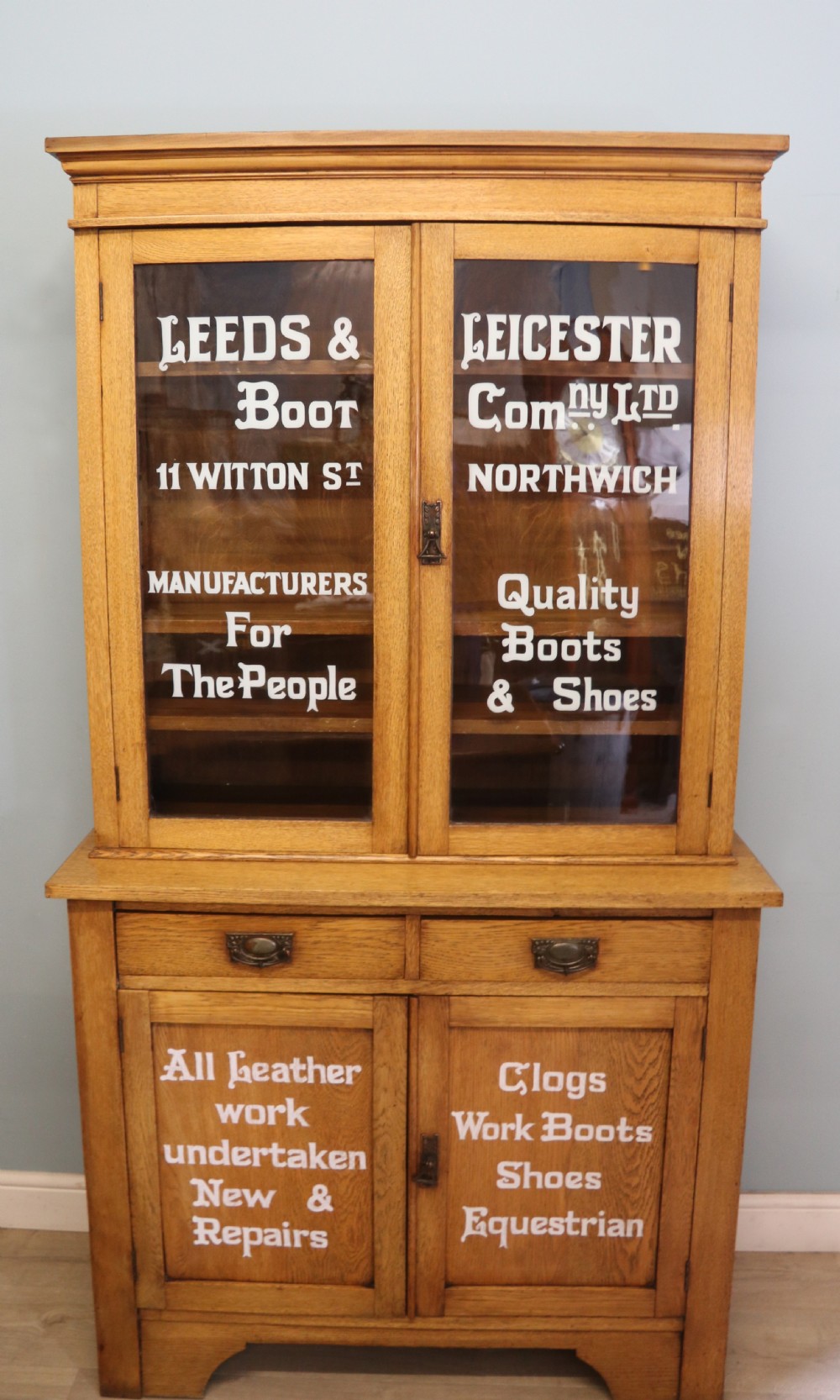 A late nineteenth-century light oak display cabinet featuring glazed upper doors and panelled lower doors decorated with hand-painted white commercial signage.