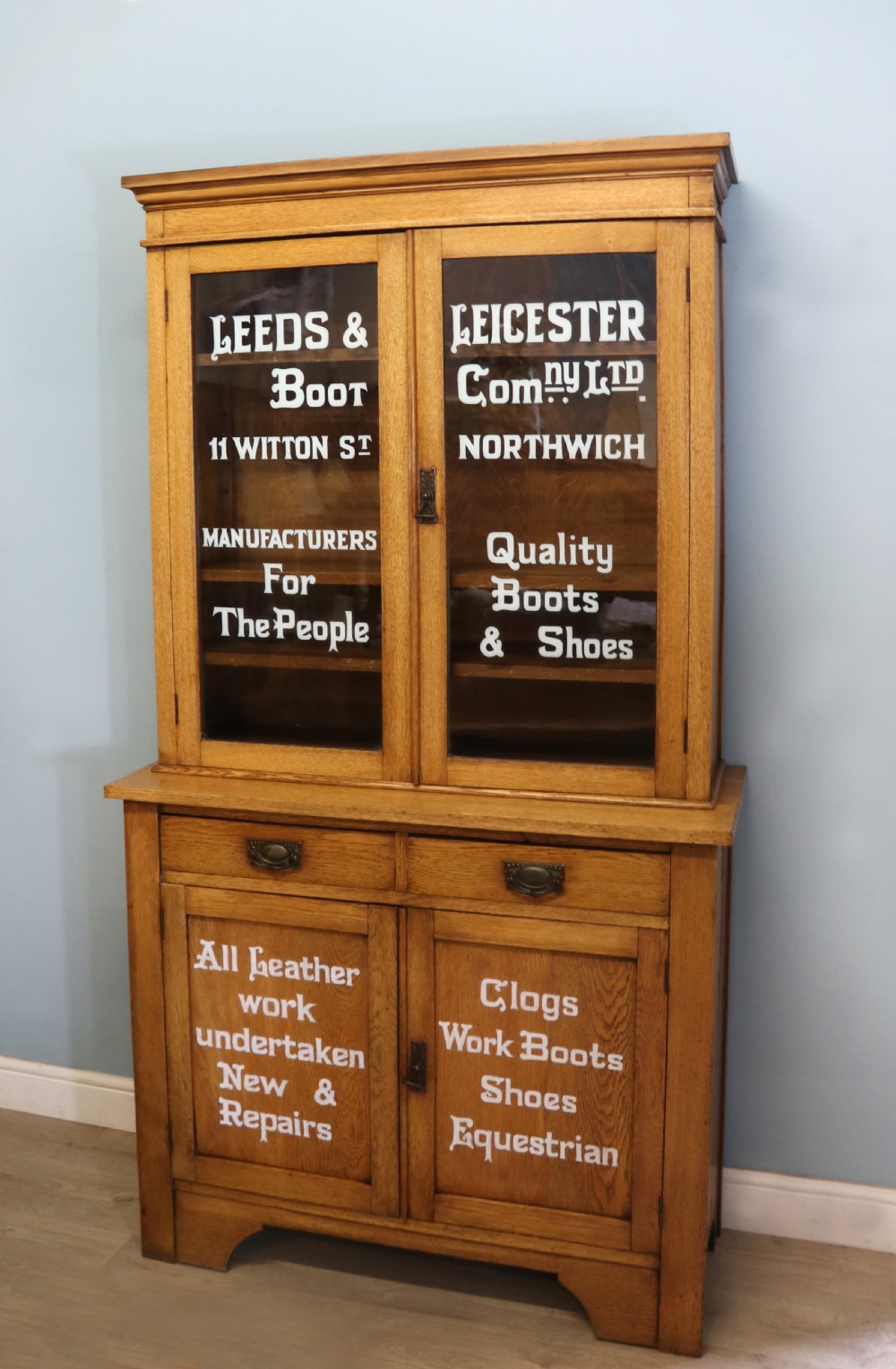 A late nineteenth-century light oak display cabinet featuring glazed upper doors and panelled lower doors decorated with hand-painted white commercial signage. View II