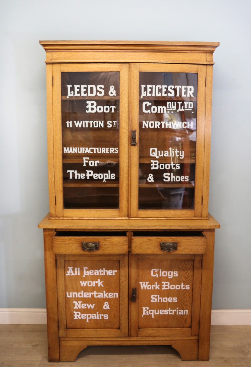 A late nineteenth-century light oak display cabinet featuring glazed upper doors and panelled lower doors decorated with hand-painted white commercial signage. View III