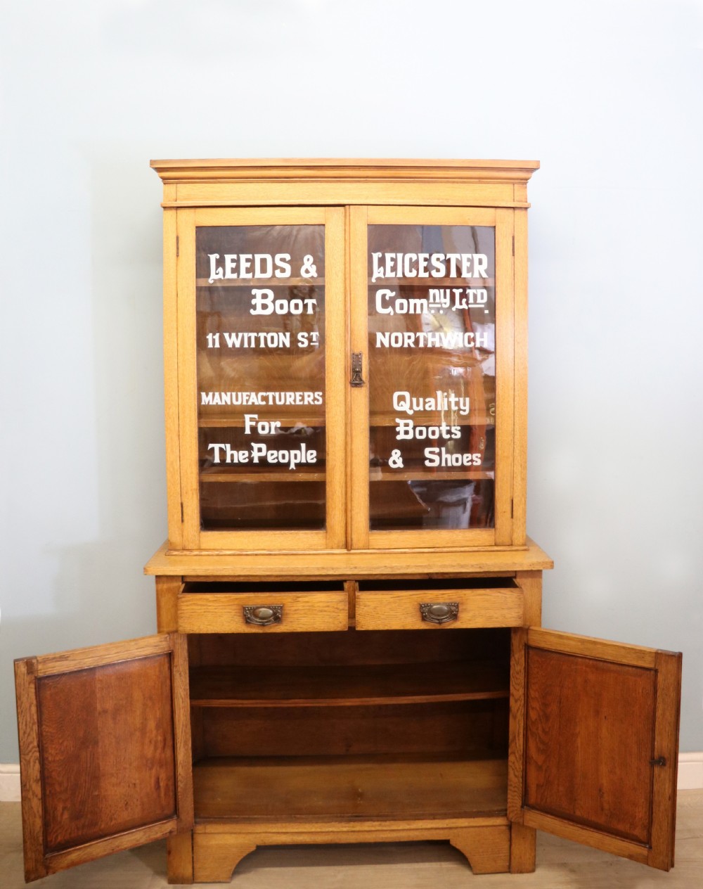 A late nineteenth-century light oak display cabinet featuring glazed upper doors and panelled lower doors decorated with hand-painted white commercial signage. View IV