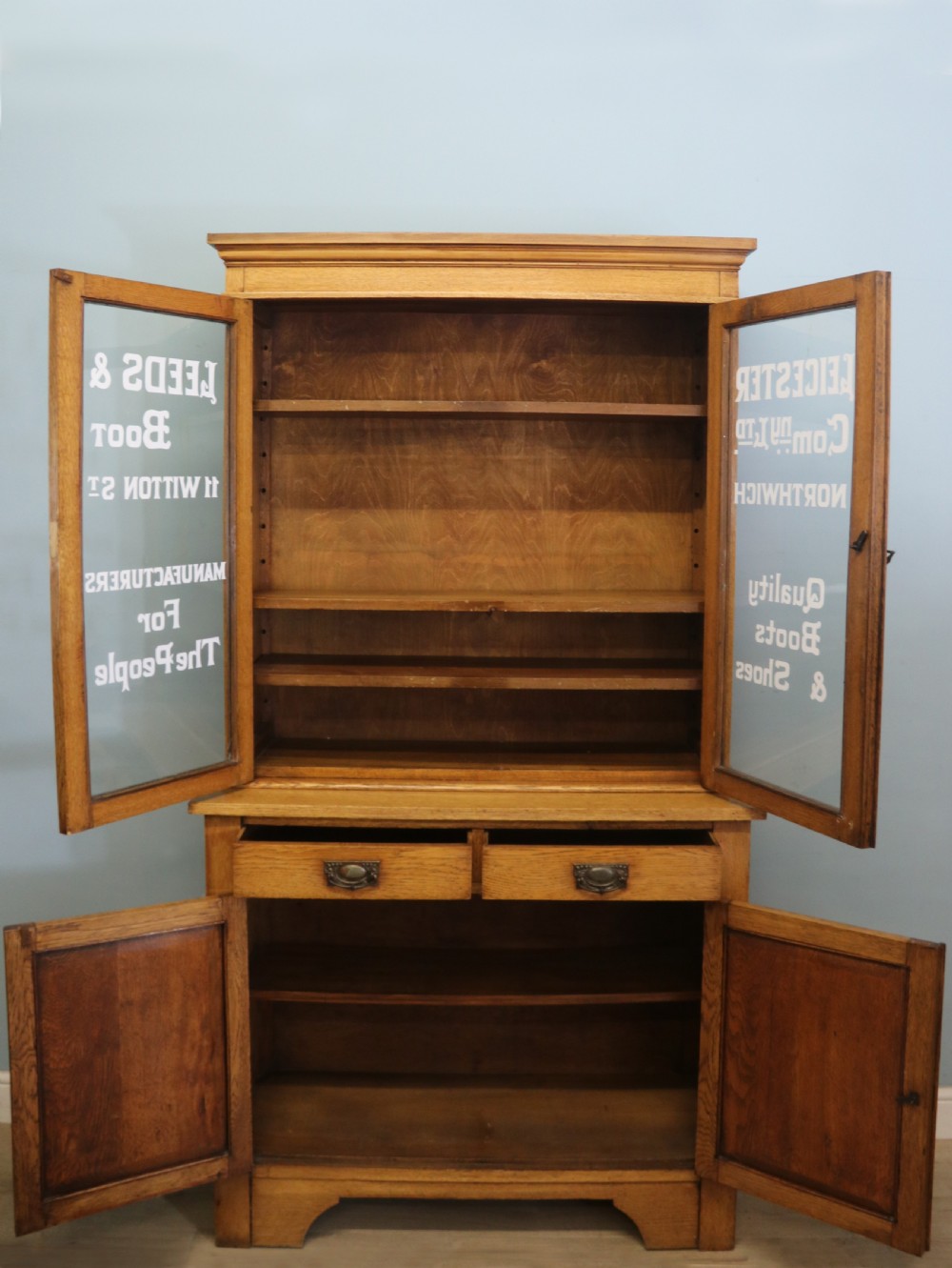 A late nineteenth-century light oak display cabinet featuring glazed upper doors and panelled lower doors decorated with hand-painted white commercial signage. View V