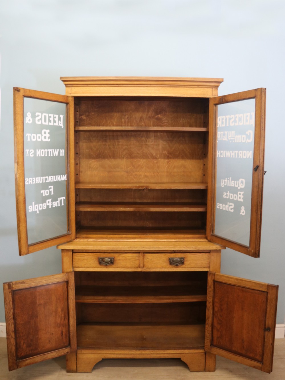 A late nineteenth-century light oak display cabinet featuring glazed upper doors and panelled lower doors decorated with hand-painted white commercial signage. View VII