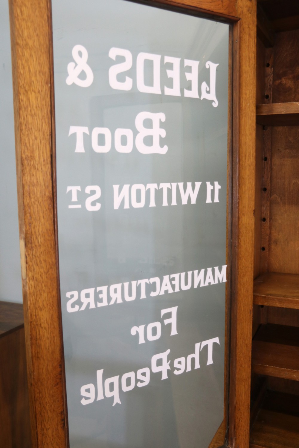 A late nineteenth-century light oak display cabinet featuring glazed upper doors and panelled lower doors decorated with hand-painted white commercial signage. View XII