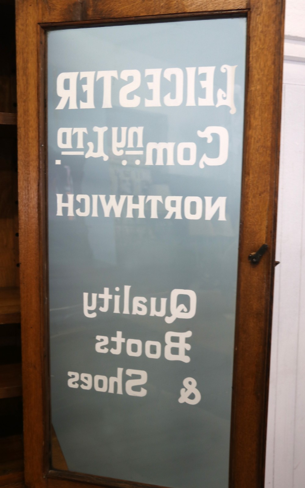 A late nineteenth-century light oak display cabinet featuring glazed upper doors and panelled lower doors decorated with hand-painted white commercial signage. View XIII