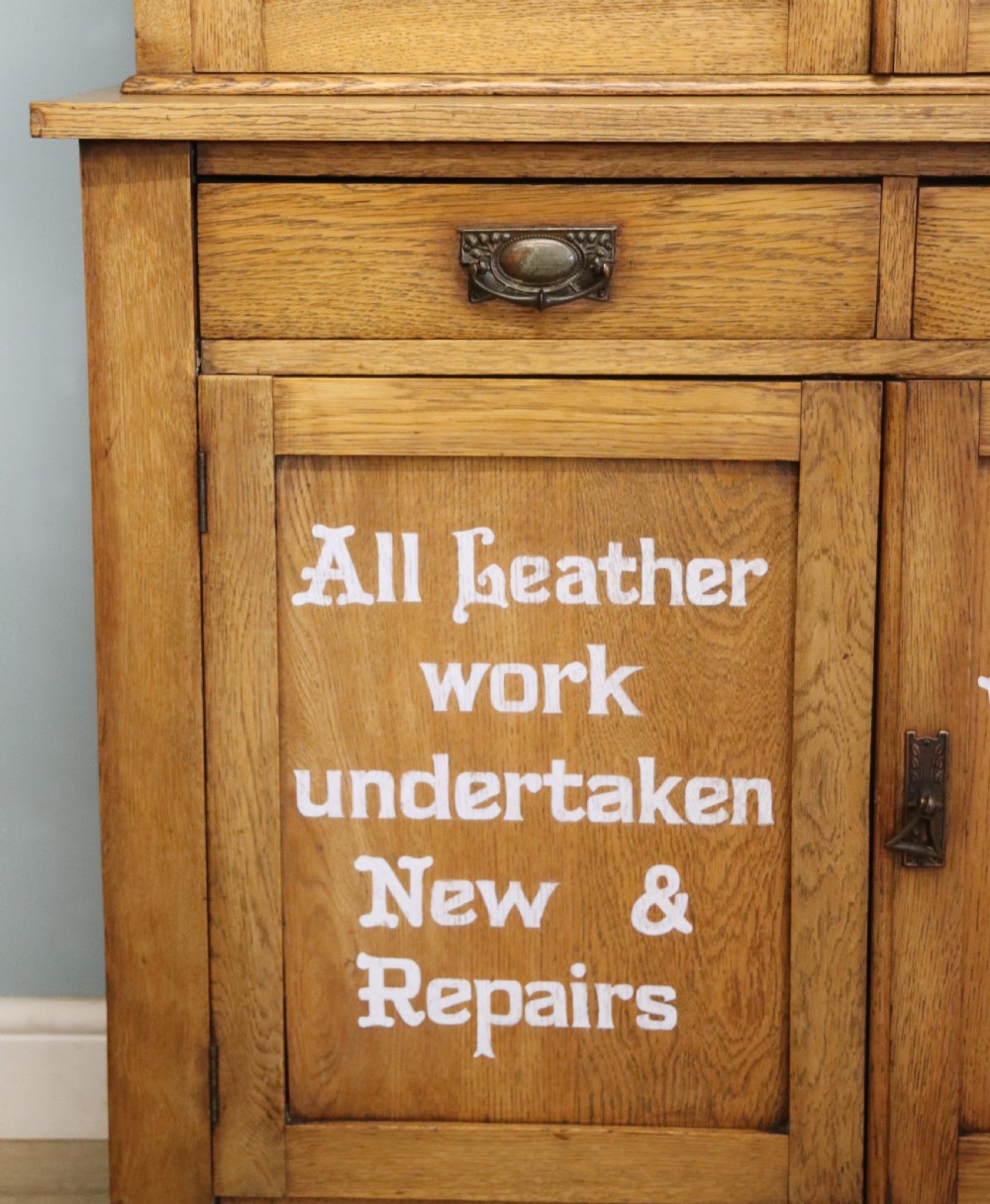 A late nineteenth-century light oak display cabinet featuring glazed upper doors and panelled lower doors decorated with hand-painted white commercial signage. View XX
