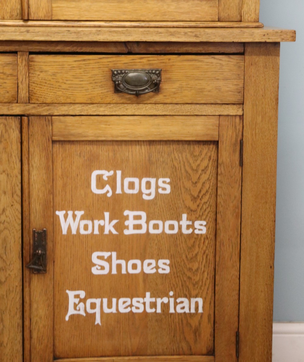 A late nineteenth-century light oak display cabinet featuring glazed upper doors and panelled lower doors decorated with hand-painted white commercial signage. View XXI