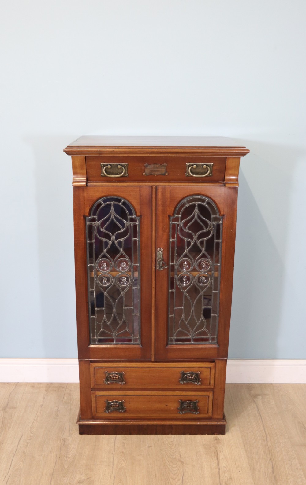 An early 20th-century walnut side cabinet featuring arched leaded glass doors, a top drawer, and two lower drawers with metal handles.