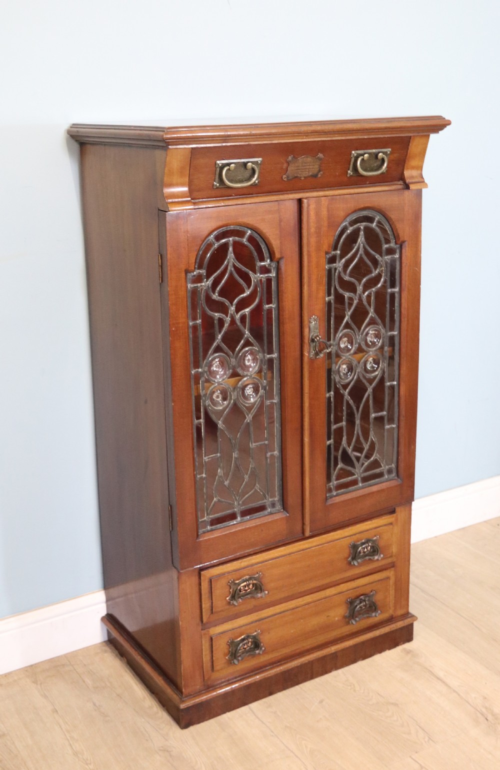 An early 20th-century walnut side cabinet featuring arched leaded glass doors, a top drawer, and two lower drawers with metal handles. View II