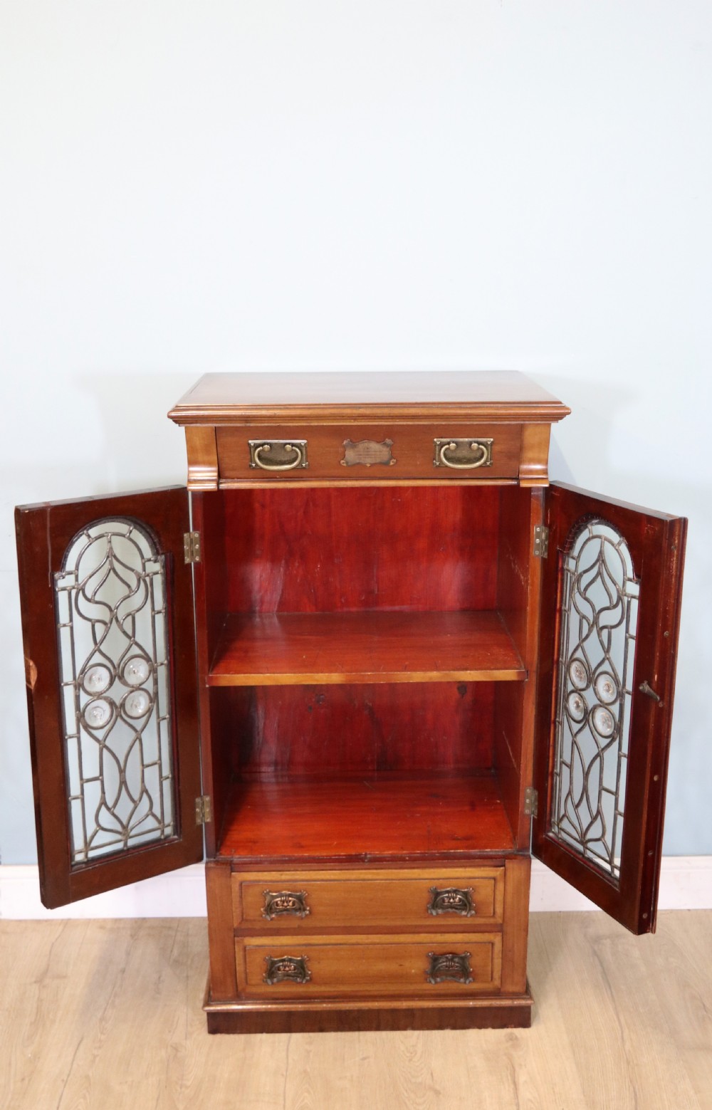 An early 20th-century walnut side cabinet featuring arched leaded glass doors, a top drawer, and two lower drawers with metal handles. View IV