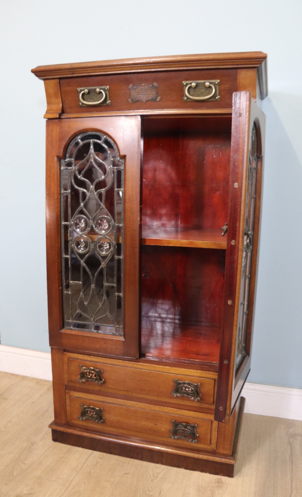 An early 20th-century walnut side cabinet featuring arched leaded glass doors, a top drawer, and two lower drawers with metal handles. View V