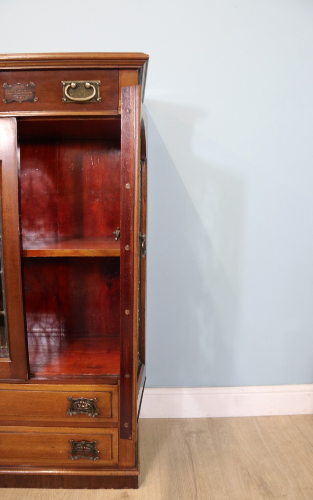 An early 20th-century walnut side cabinet featuring arched leaded glass doors, a top drawer, and two lower drawers with metal handles. View VI