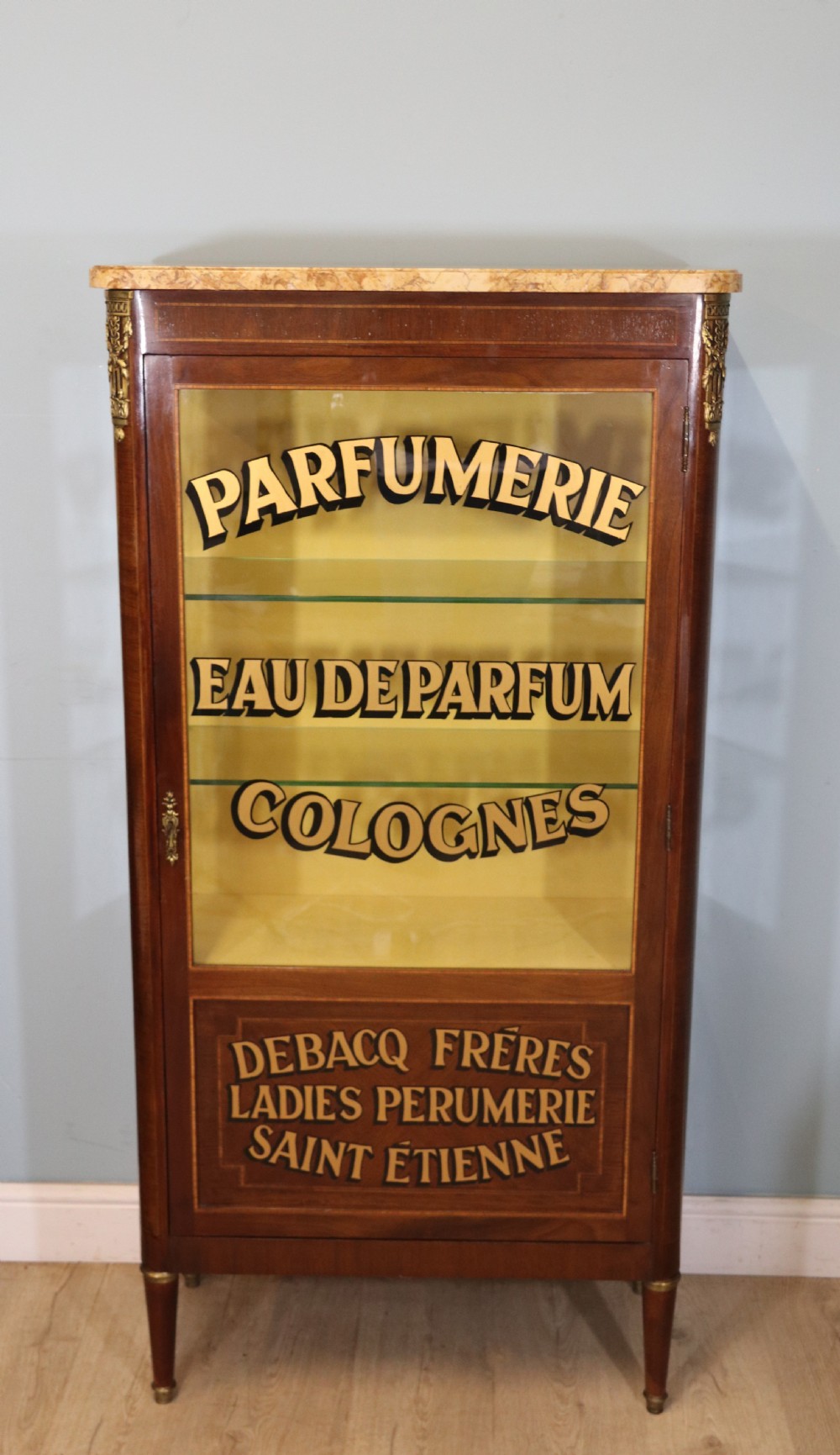 A French mahogany vitrine featuring a marble top, glass display door with gilt perfumery lettering, glass shelves, and brass mounts.