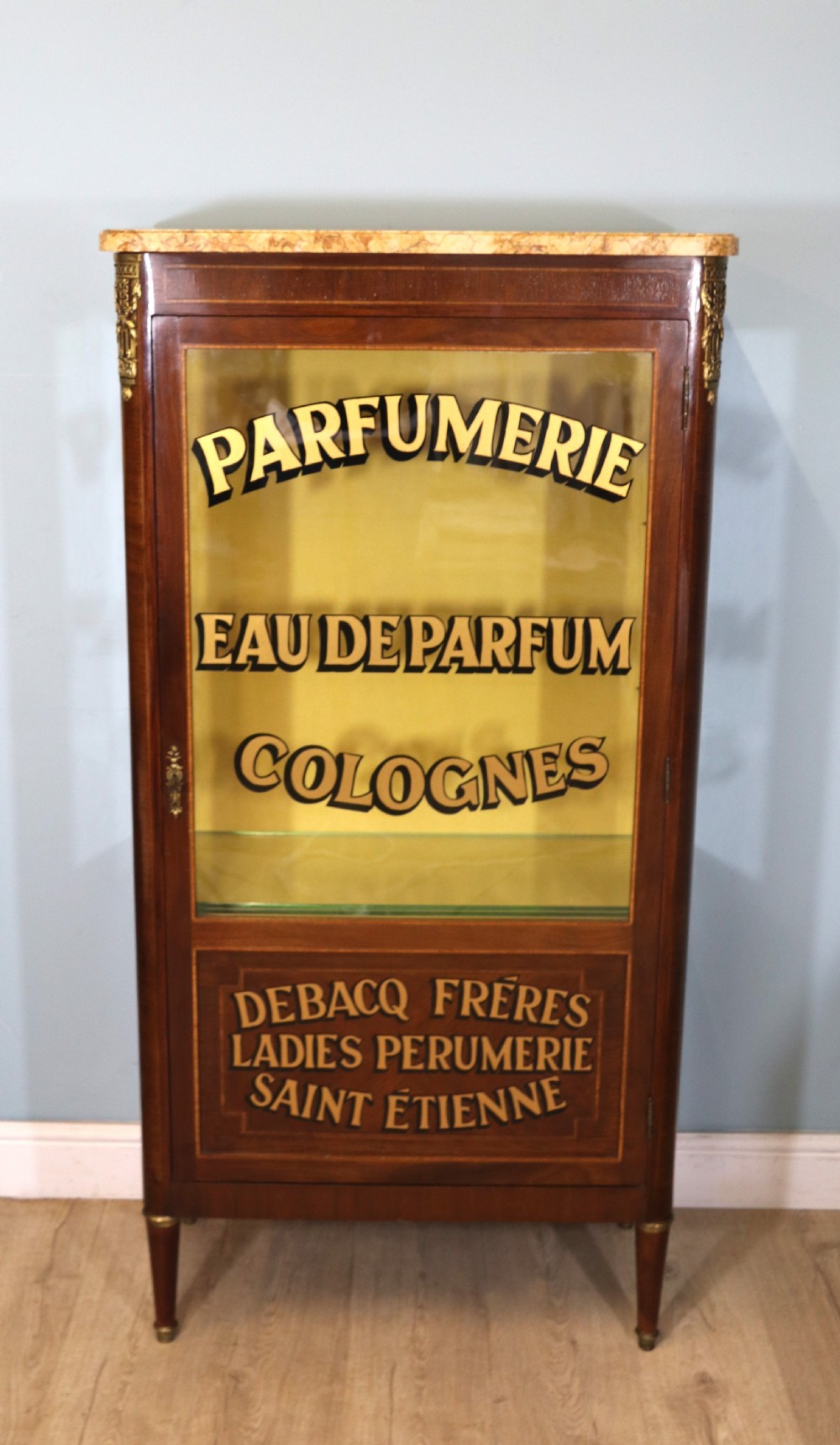 A French mahogany vitrine featuring a marble top, glass display door with gilt perfumery lettering, glass shelves, and brass mounts. View II