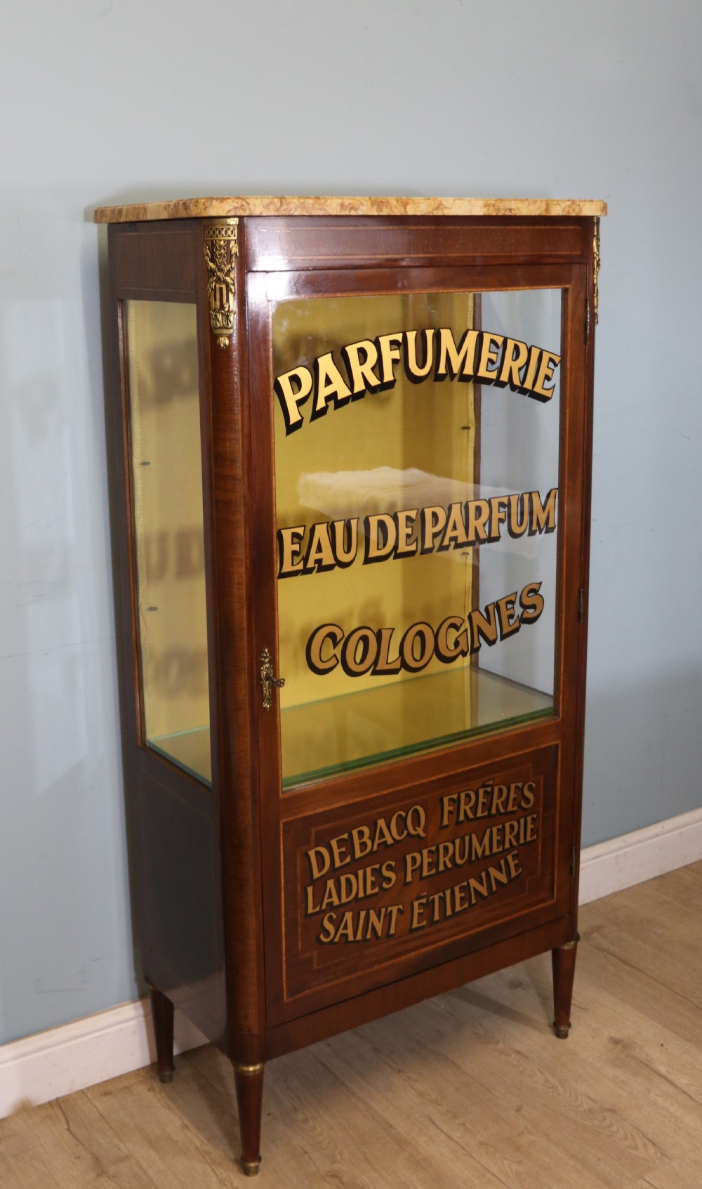 A French mahogany vitrine featuring a marble top, glass display door with gilt perfumery lettering, glass shelves, and brass mounts. View IV