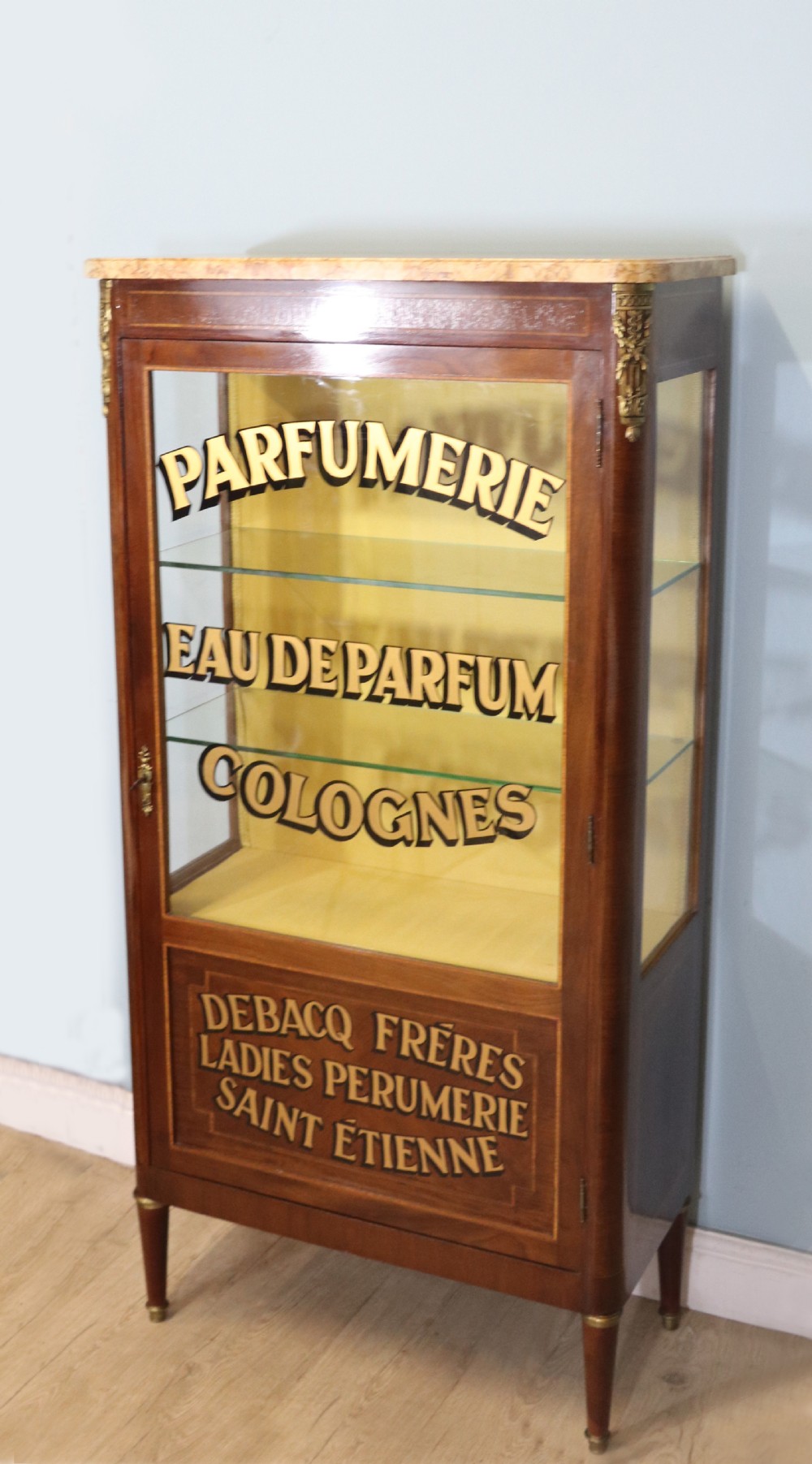 A French mahogany vitrine featuring a marble top, glass display door with gilt perfumery lettering, glass shelves, and brass mounts. View V