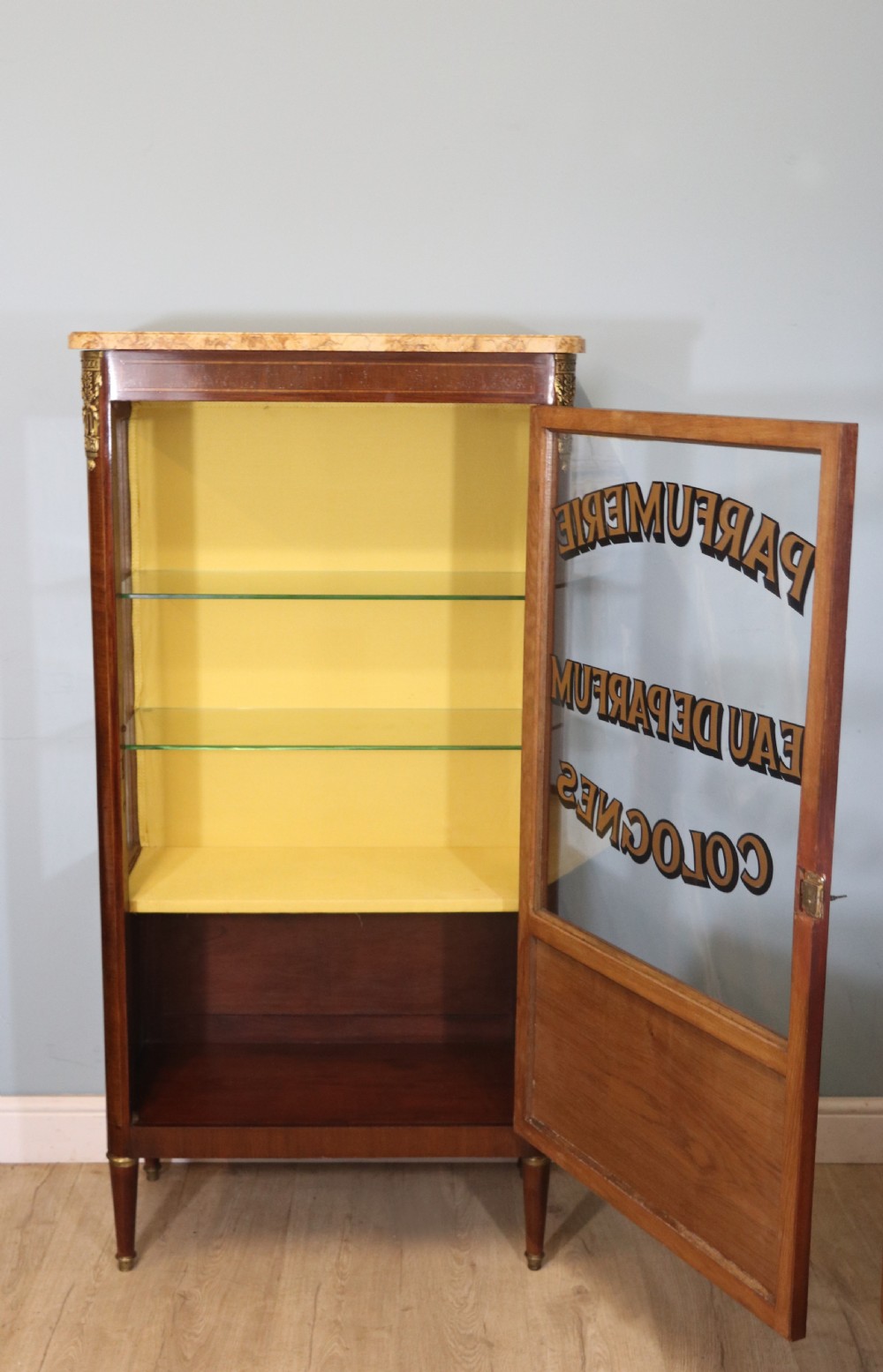 A French mahogany vitrine featuring a marble top, glass display door with gilt perfumery lettering, glass shelves, and brass mounts. View VI