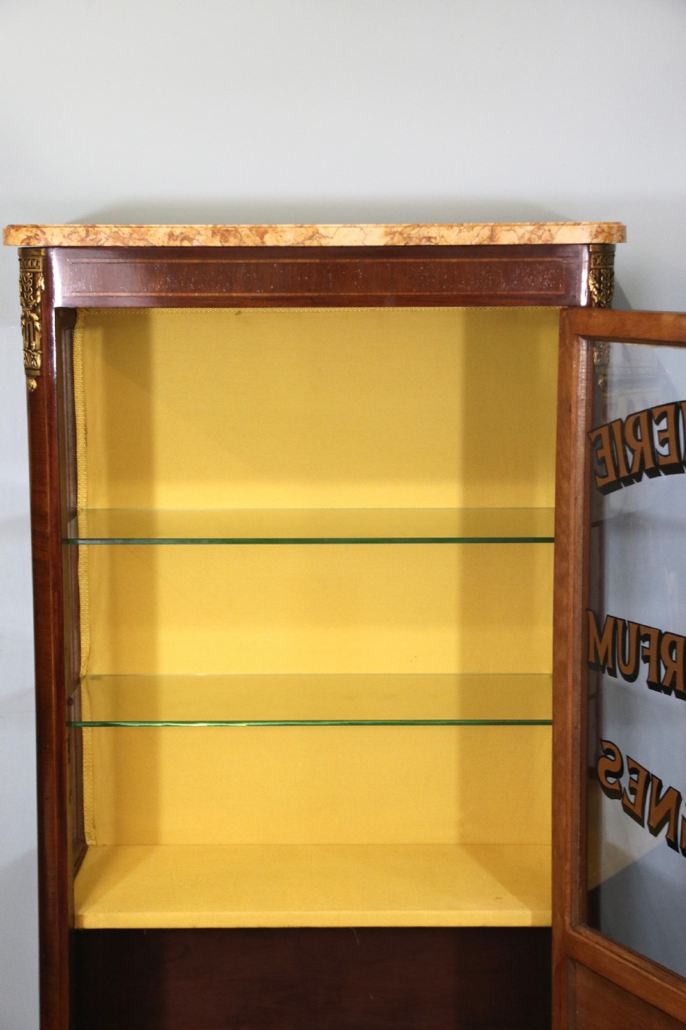 A French mahogany vitrine featuring a marble top, glass display door with gilt perfumery lettering, glass shelves, and brass mounts. View XII