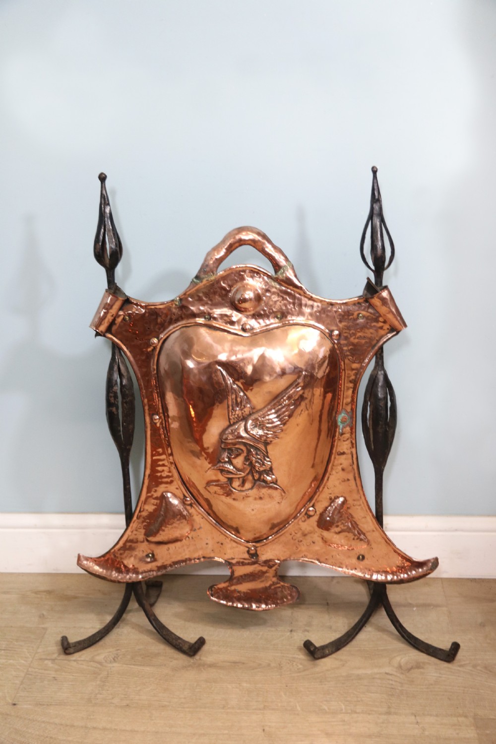 A 19th-century repousse copper panel fire screen featuring a winged warrior profile motif mounted in a wrought-iron frame with splayed feet.