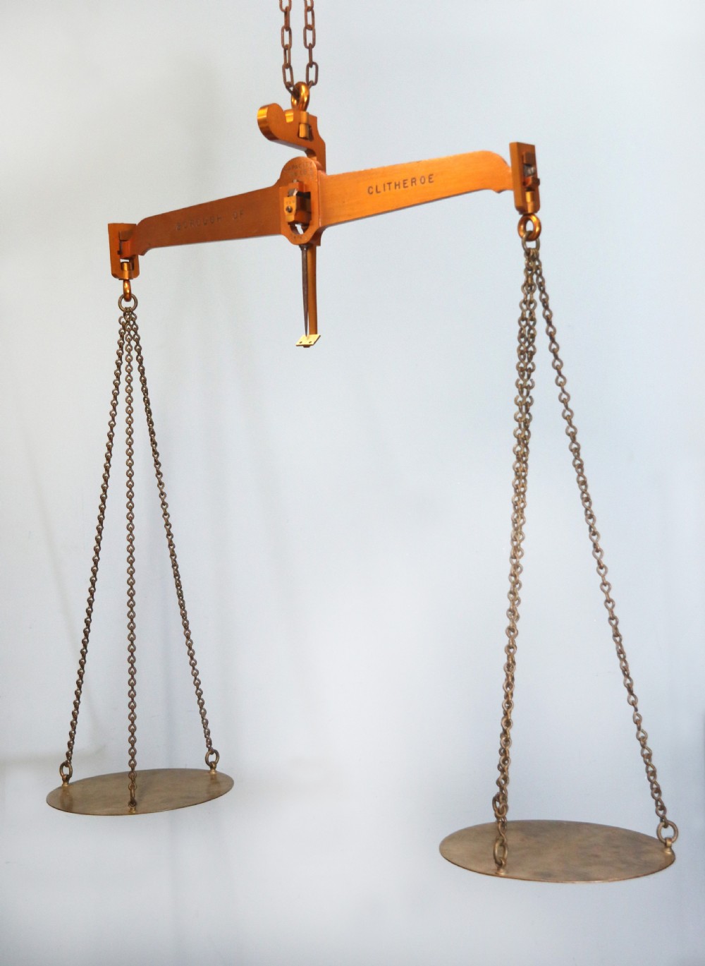 A large hanging brass beam balance scale featuring stamped lettering, suspended chain links, and two circular brass pans.