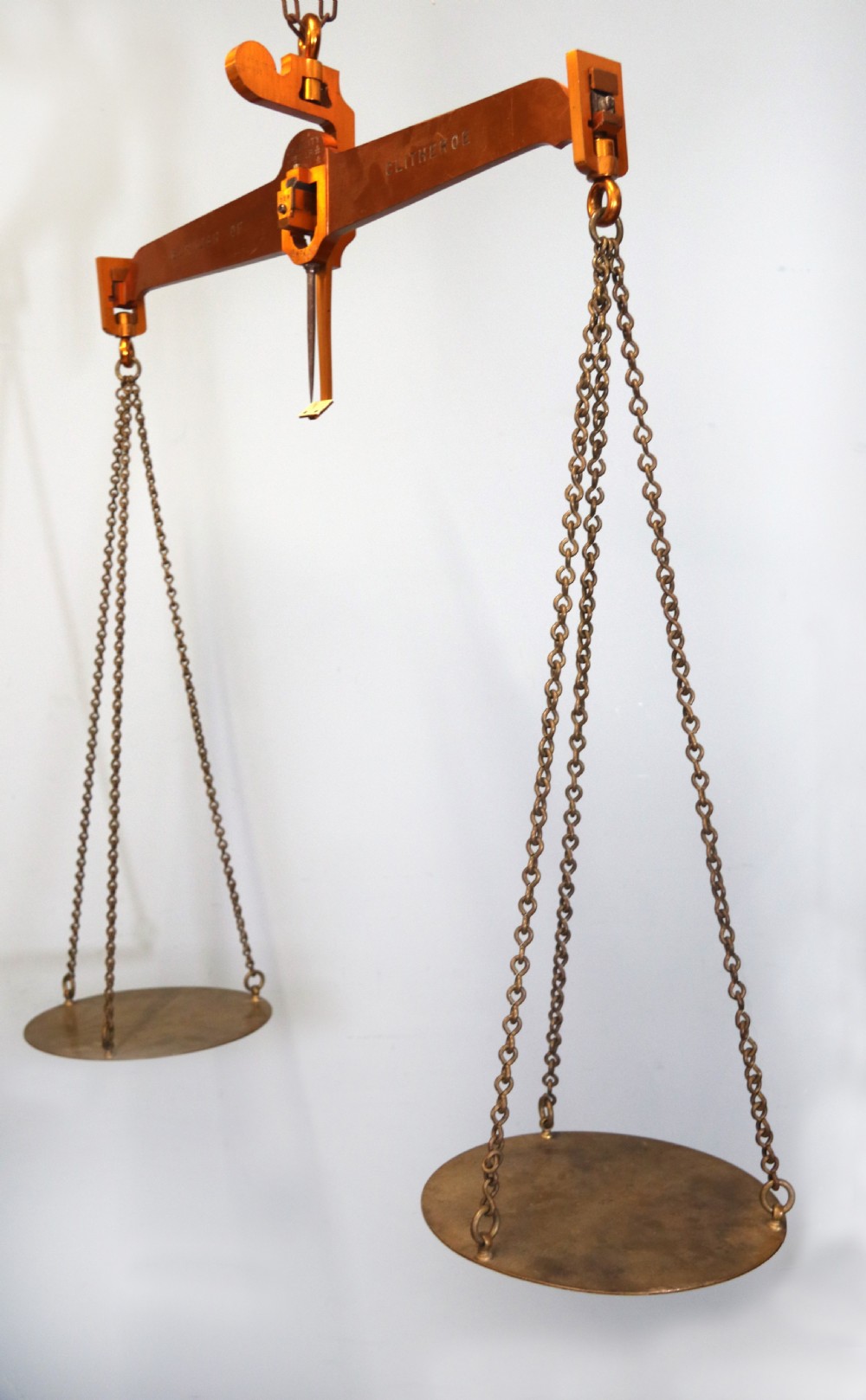 A large hanging brass beam balance scale featuring stamped lettering, suspended chain links, and two circular brass pans. View II