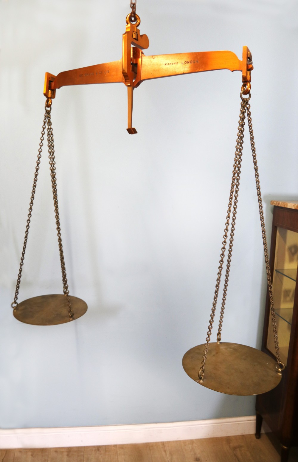 A large hanging brass beam balance scale featuring stamped lettering, suspended chain links, and two circular brass pans. View III