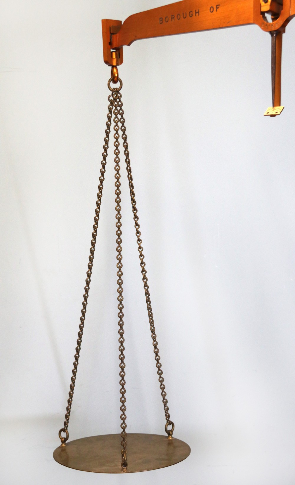 A large hanging brass beam balance scale featuring stamped lettering, suspended chain links, and two circular brass pans. View V